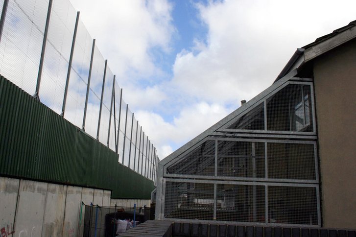 Die „Friedenslinie“ oder „Friedensmauer“ in der Bombay Street in Belfast im Mai 2009. Die kleinen Hintergärten der Häuser mussten durch Metallkäfige geschützt werden, da Geschosse häufig über die Mauer geworfen wurden. (Foto: Supermac1961, CC BY 2.0)