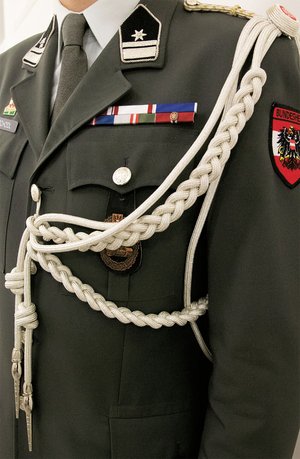 Das Verwendungsabzeichen Schulterschnur Attachéunteroffizier, mattsilber, wird von den AttUO vorschriftsmäßig an der linken Schulter getragen und besteht aus einer vierteiligen Grundschnur, wovon zwei Teile jeweils zu Zöpfen mit 36 bzw. 20 Rapporten geflochten sind und in einer silbernen Metallspitze münden. Die Schnurverbindung selbst erfolgt über einen Blechaufsatz, der mit einer mit dem Staatswappen ausgeführten Kokarde verdeckt wird und zur Befestigung an der Schulterspange dient. (Foto: RedTD/Miriam Gamauf)