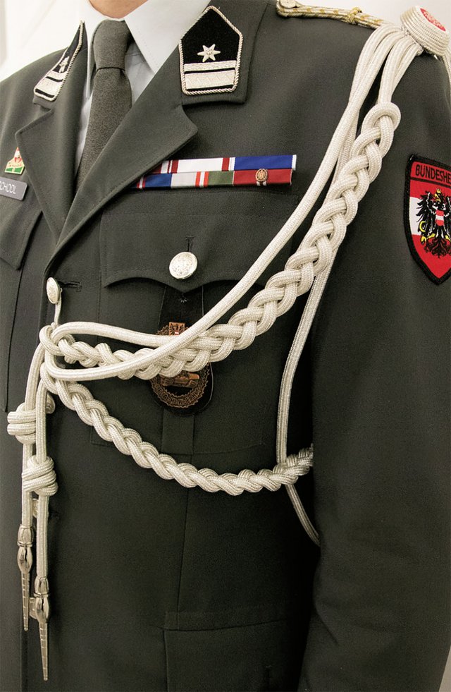 Das Verwendungsabzeichen Schulterschnur Attachéunteroffizier, mattsilber, wird von den AttUO vorschriftsmäßig an der linken Schulter getragen und besteht aus einer vierteiligen Grundschnur, wovon zwei Teile jeweils zu Zöpfen mit 36 bzw. 20 Rapporten geflochten sind und in einer silbernen Metallspitze münden. Die Schnurverbindung selbst erfolgt über einen Blechaufsatz, der mit einer mit dem Staatswappen ausgeführten Kokarde verdeckt wird und zur Befestigung an der Schulterspange dient. (Foto: RedTD/Miriam Gamauf)