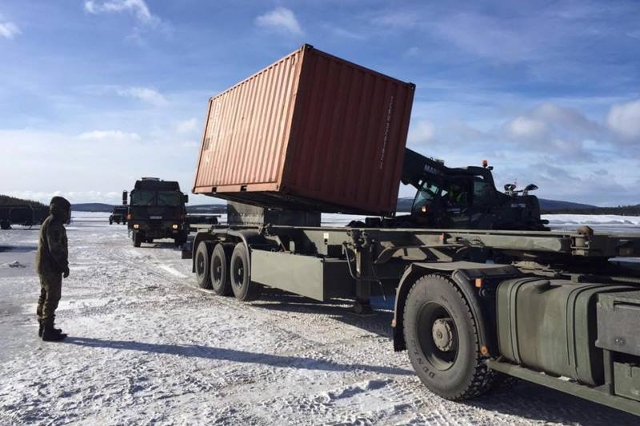 Ein LKW wird mit einem Container beladen. Links im Bild ist ein Soldat zu sehen, im Hintergrund sieht man weitere LKWs.